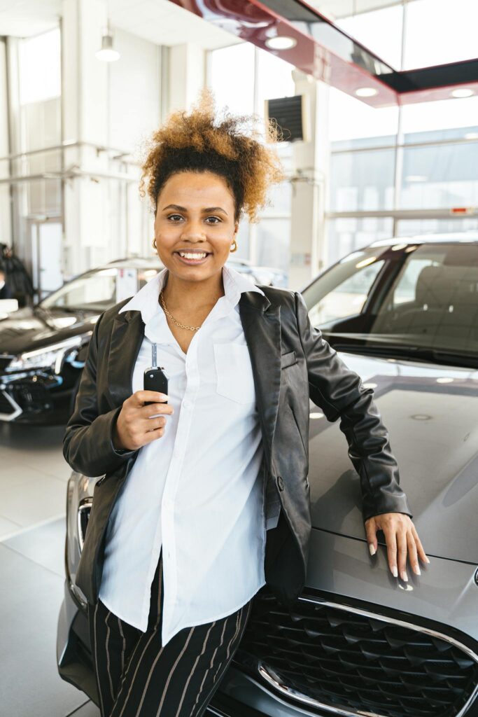 Portrait of a smiling woman holding car key next to a new car inside a dealership.